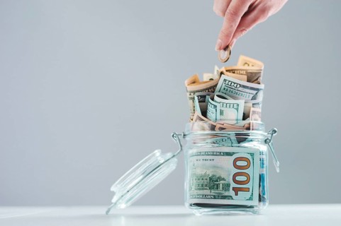 Female hand puts a coin into a glass piggy bank full of money. Thrift concept.