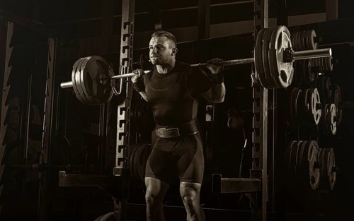 The weightlifter stands with a very heavy barbell on his shoulders. He is at the top and is preparing to sit down