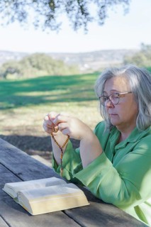 Mature white-haired woman with glasses praying with a rosary beads and a bible on a wooden table in the field