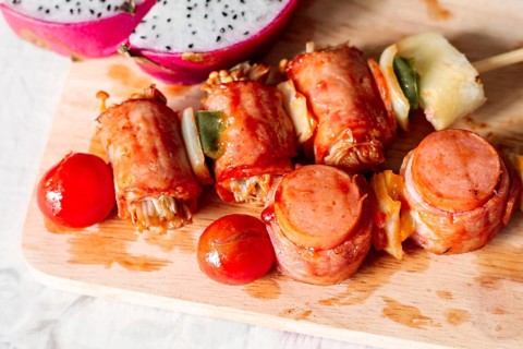 Skewers with bacon-wrapped sausages and vegetables on a wooden cutting board with dragon fruit