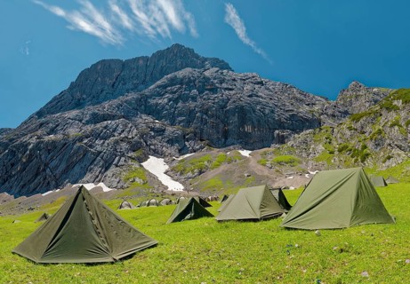 Tents of mountain hunters on a green meadow in front of an imposing mountain backdrop under a clear blue sky