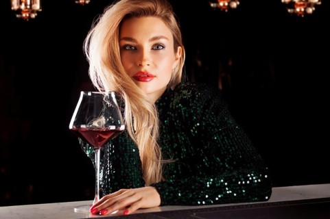 Seductive blonde in a green evening dress posing with a glass of red wine.