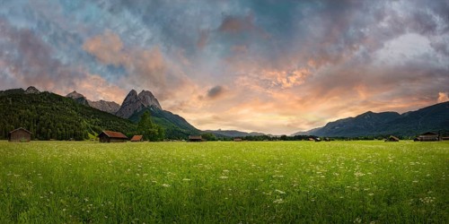 Panoramic view of a green meadow in front of imposing mountains under a cloudy evening sky