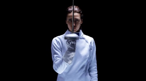 Charming girl in a fencer costume posing on a black background. The sword divides the face into two parts. The concept of fencing.