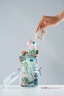 Female hand puts a bill into a glass piggy bank full of money. Thrift concept.