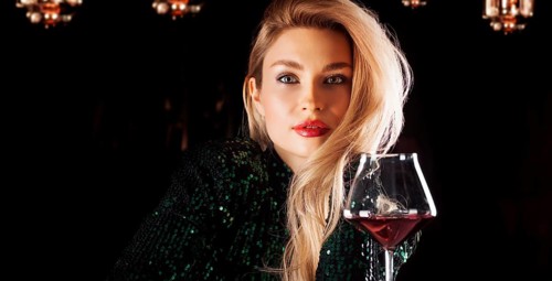 Seductive blonde in a green evening dress posing with a glass of red wine.