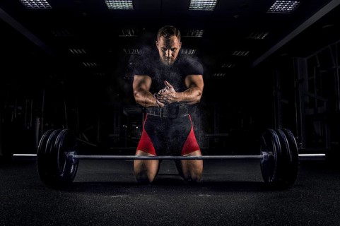 Ahlete kneels in the gym before the bar and claps his hands after which the dust from the chalk flies