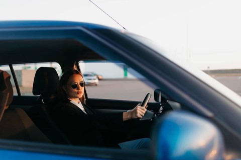 A young beautiful stylish girl driver in a jacket and sunglasses at the wheel of a car is waiting for passengers