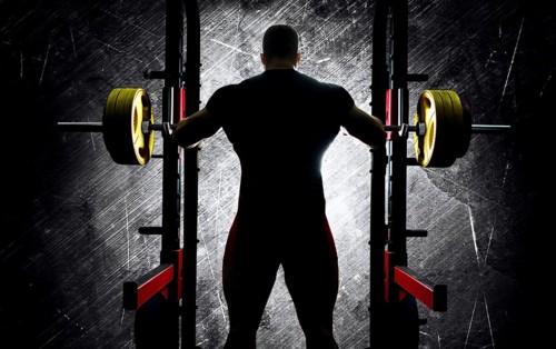 The weightlifter stands with his hands behind the heavy barbell. Back view