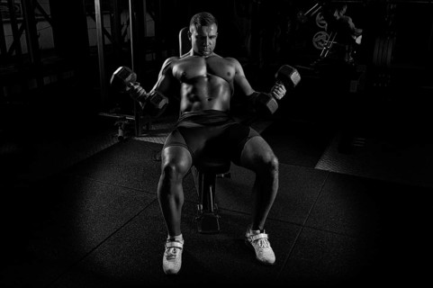 Professional athlete makes an exercise on the biceps by lifting dumbbells while sitting on the bench. View from above