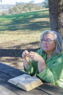 Older white-haired woman with a rosary beads in her hand and a bible praying with her eyes closed sitting at a wooden table in the forest