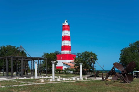 Red and white lighthouse under a blue sky next to green areas and a historic anchor