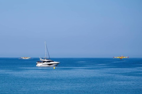 Two fire-fighting aircraft refuelling water at the sea with sailing yacht in the foreground