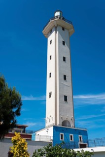 A white lighthouse with a blue base against a blue sky surrounded by green and yellow