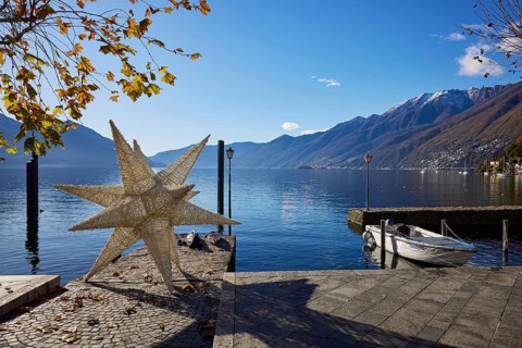 Backlit photograph of the large Herrenhuth star as a Christmas decoration at the harbour entrance to Lake Maggiore with lanterns and motorboat in Ascona