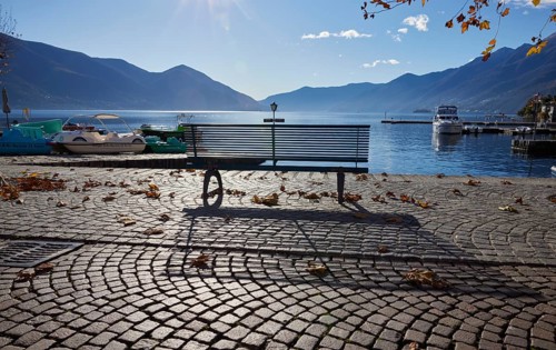 Backlit photograph of a public bench with shade and lake view on the Lungolago