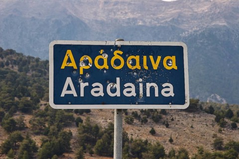 Damaged road sign with Greek and Latin lettering in front of a mountain landscape, Aradena Gorge,