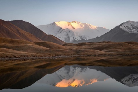White glaciated and snowy mountain peak Pik Lenin at sunset, mountains reflected in a lake between