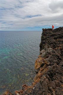 A brave person stands on the edge of a high volcanic cliff above the sea, lava rocks coastal hiking