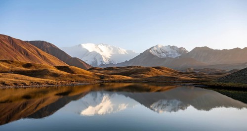 White glaciated and snowy mountain peak Pik Lenin at sunset, mountains reflected in a lake between