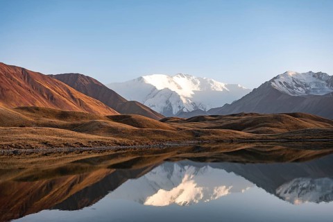 White glaciated and snowy mountain peak Pik Lenin at sunset, mountains reflected in a lake between