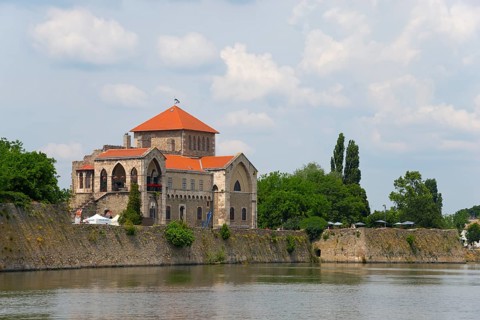 A historic building stands by a quiet river, surrounded by green nature and a light cloudy sky,