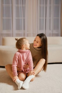 A young mother and daughter sit on the floor in a cozy living room, with a couch and curtains in