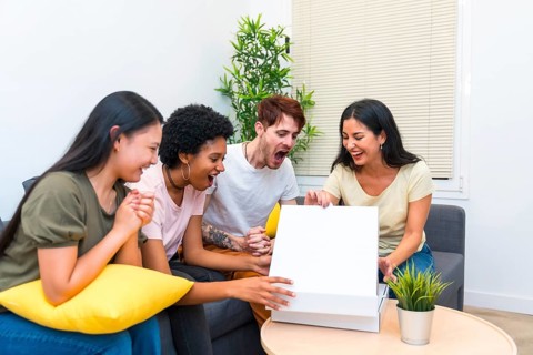 Happy multiracial friends sitting on a couch opening pizza box to eat at home