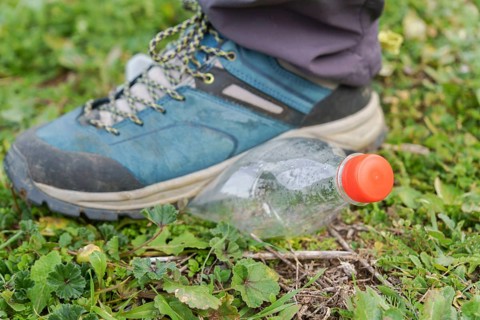 Hiking boot stepping on an empty plastic bottle on the green grass of a meadow, ecology and