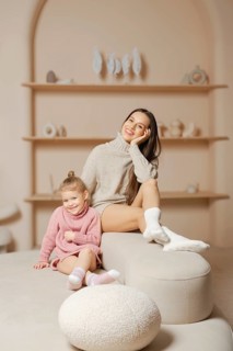 Happy young mother in warm sweater and socks sitting on top of a white ottoman next to her daughter