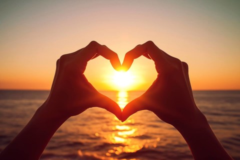 Two hands forming a heart shape, silhouetted against a breathtaking ocean sunset. Love and warmth