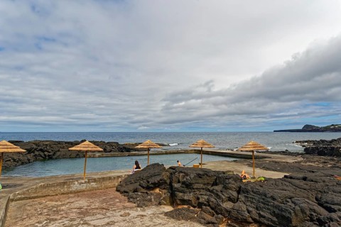 Natural swimming pool by the sea with parasols and people relaxing, lava rocks coastal walk Ponta