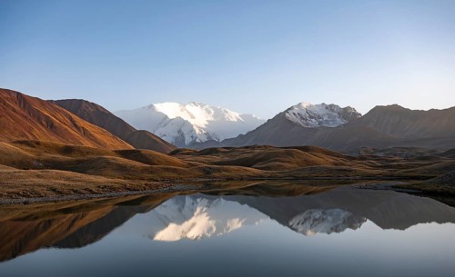 White glaciated and snowy mountain peak Pik Lenin at sunset, mountains reflected in a lake between