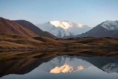 White glaciated and snowy mountain peak Pik Lenin at sunset, mountains reflected in a lake between