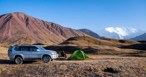 An off-road vehicle stands next to a pitched tent in a mountain landscape with snow-capped peaks in