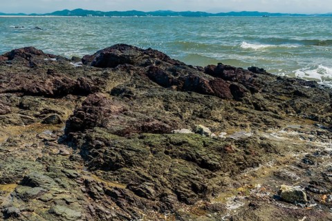 Seascape of rocking shore with beach and harbor road in background under cloudy blue sky