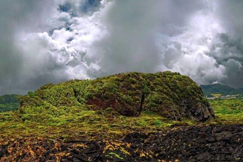 Green hills and dark volcanic rocks under a dramatic cloudy sky, lava rocks coastal hiking trail