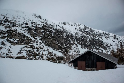 A solitary cabin sits beneath a rugged, snowy mountain landscape under an overcast sky. Poland,
