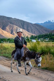 Man in traditional dress riding a donkey in front of a mountain backdrop, Kyrgyzstan, Asia