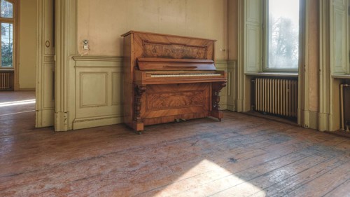 An old piano in a room with a wooden floor and large windows through which sunlight falls, Villa