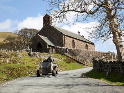 Farmer driving quad bike past village church, Buttermere, Cumbria, England, UK