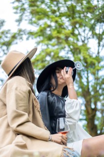 Lifestyle of young friends in hats sitting on the grass toasting with wine. Side view of two women