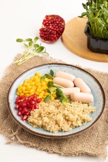 Mixed quinoa porridge, sweet corn, pomegranate seeds and small sausages on white wooden background.
