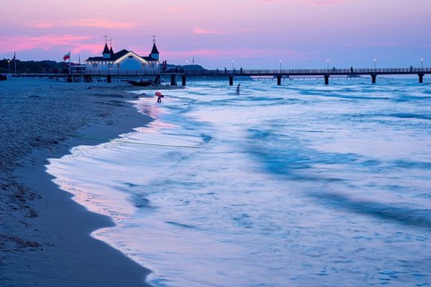 Calm evening mood on the beach with calm waves and two people near a pier with a building at dusk,