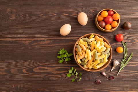 Rigatoni colored raw pasta with tomato, eggs, spices, herbs on brown wooden background. Top view,