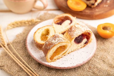 Homemade sweet bun with apricot jam and cup of coffee on white wooden background and linen textile.