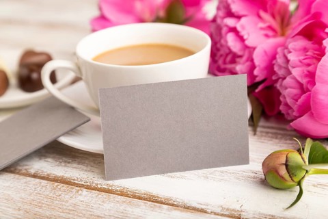 Gray business card with pink peony flowers and cup of coffee on white wooden background. side view,