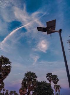 A silhouette of a solar-powered streetlamp against evening sky with clouds and palm trees,