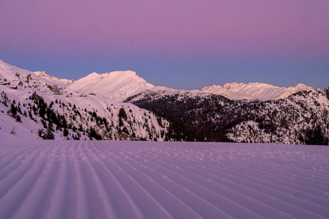 Snow-covered mountains at dusk with a purple sky and a freshly groomed ski slope in the foreground,