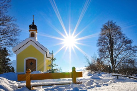 Backlit photograph of the snow-covered Maria Chapel on the hummocky meadows of Werdenfelser Land